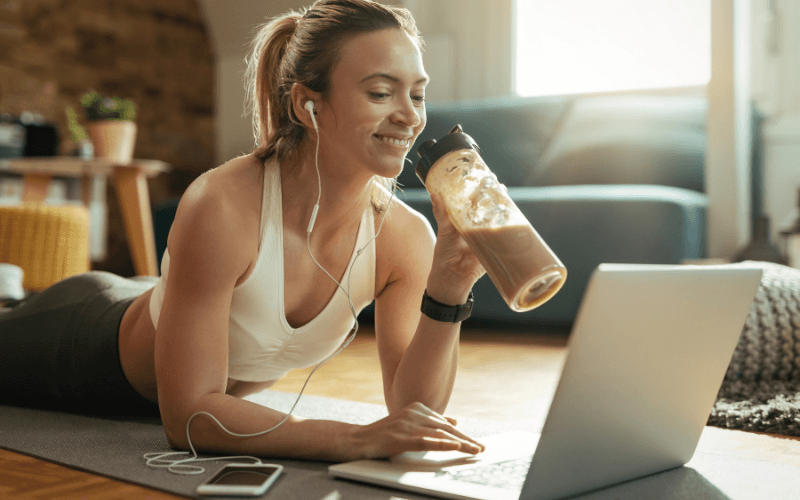 Mulher em casa sorrindo ao notebook e tomando um shake de proteína para prevenir a relação entre mounjaro e queda de cabelo.