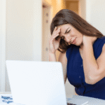 Mulher sentada em frente ao notebook com a mão na cabeça demonstrando sentir cansaço constante e exaustão.