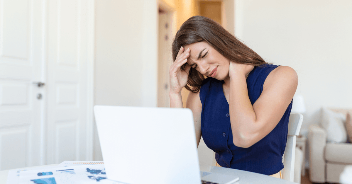 Mulher sentada em frente ao notebook com a mão na cabeça demonstrando sentir cansaço constante e exaustão.